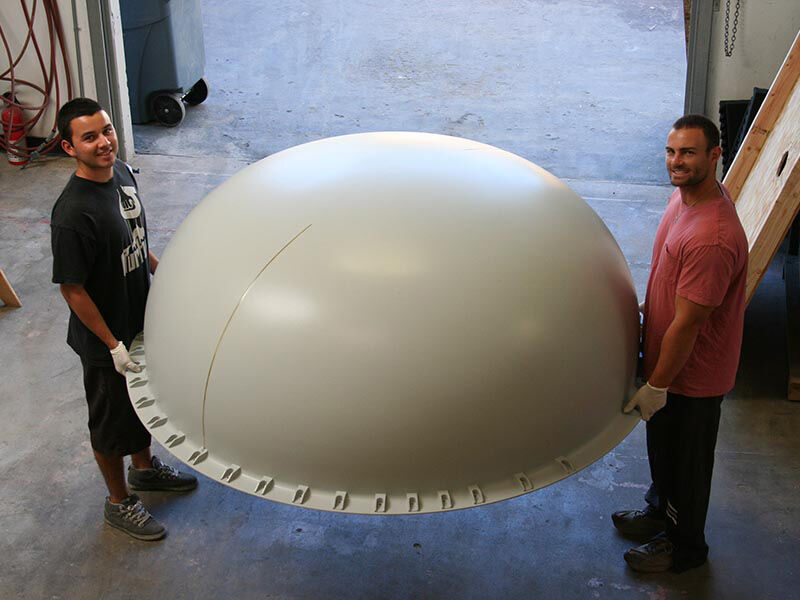 Two technicians holding up the very large and completed Ku-band belly radome in a manufacturing bay. 
